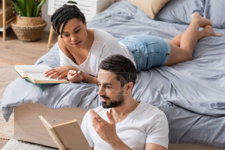 bearded man gesturing and reading book to african american girlfriend lying on bed at home