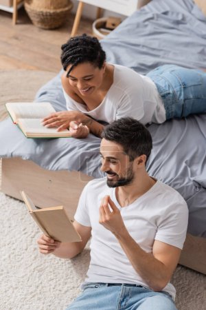 high angle view of cheerful bearded man reading book to smiling african american woman lying on bed at home