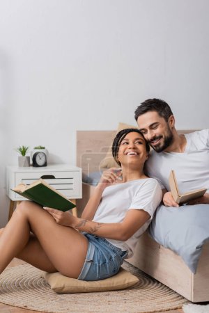joyeuse femme afro-américaine avec livre assis sur le sol et oreiller près de copain barbu couché sur le lit à la maison