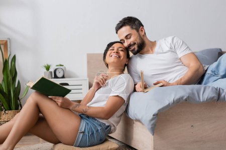 carefree african american woman with book smiling with closed eyes while sitting near smiling boyfriend lying on bed at home