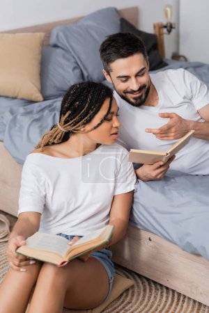 homme barbu souriant pointant vers livre près de jeune femme afro-américaine dans la chambre à coucher à la maison