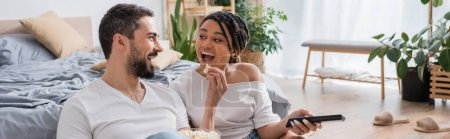 excited bearded man feeding african american woman with popcorn while watching tv in bedroom at home, banner
