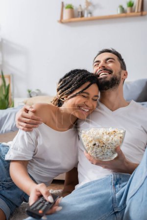 laughing bearded man with popcorn hugging smiling african american woman with tv remote controller in bedroom at home