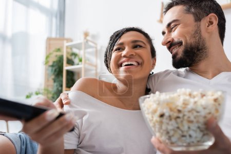 homme barbu souriant avec bol de maïs soufflé étreignant femme afro-américaine insouciante en cliquant sur les chaînes de télévision dans la chambre à coucher à la maison