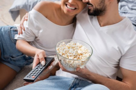 Teilansicht eines fröhlichen gemischtrassigen Paares mit Schüssel Popcorn und Fernbedienung im heimischen Schlafzimmer