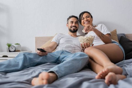 pleased african american woman eating popcorn near smiling bearded man with tv remote controller on bed at home