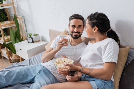 cheerful interracial couple with tv remote controller and popcorn smiling at each other on bed at home
