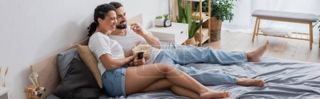 barefoot and carefree interracial couple eating popcorn and watching movie on bed at home, banner