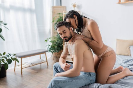 sexy african american woman in underwear seducing shirtless bearded man in jeans in modern bedroom