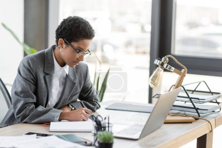 young african american businesswoman in eyeglasses looking at laptop and writing in notebook while working in office