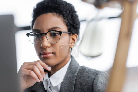portrait of african american businesswoman in grey blazer and eyeglasses holding hand near face while thinking in office