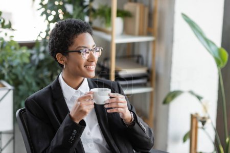 Téléchargez les photos : Joyeuse femme d'affaires afro-américaine en veston noir et lunettes tenant tasse de café et regardant loin dans le bureau - en image libre de droit