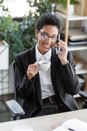 happy african american businesswoman in eyeglasses and black blazer sitting with coffee cup and talking on cellphone in office