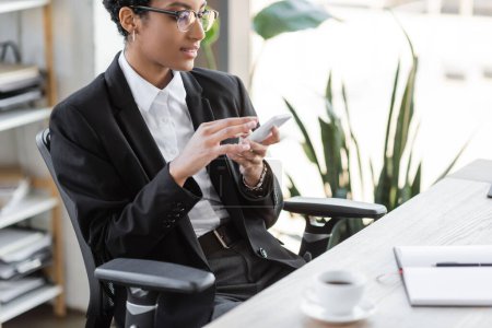 Téléchargez les photos : Femme d'affaires afro-américaine positive dans les lunettes et la messagerie blazer élégant sur téléphone mobile près de tasse de café floue dans le bureau - en image libre de droit