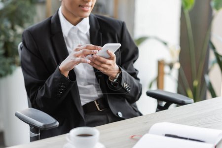 Teilbild einer afrikanisch-amerikanischen Geschäftsfrau im schwarzen Blazer, die in der Nähe einer verschwommenen Kaffeetasse im Büro auf ihrem Handy Nachrichten sendet