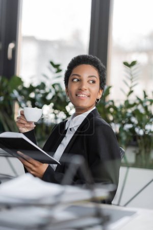 freudige afrikanisch-amerikanische Geschäftsfrau schaut weg, während sie mit Kaffeetasse und Notizbuch im Büro sitzt