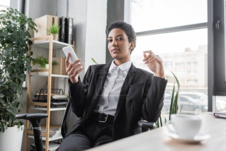 young african american businesswoman in black suit holding mobile phone and looking at camera near blurred coffee cup in office