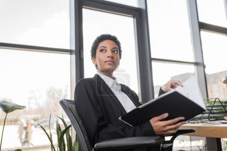 pensive african american businesswoman with folder sitting and looking away in office