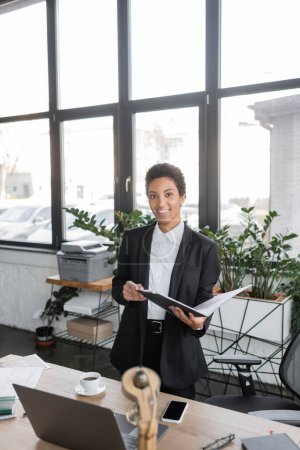 Téléchargez les photos : Heureuse femme d'affaires afro-américaine tenant dossier avec des documents et regardant la caméra tout en se tenant près de l'ordinateur portable dans le bureau moderne - en image libre de droit