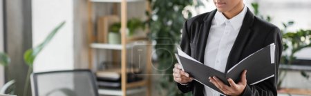 Téléchargez les photos : Vue partielle d'une femme d'affaires afro-américaine dans un blazer noir tenant un dossier avec des papiers au bureau, une bannière - en image libre de droit