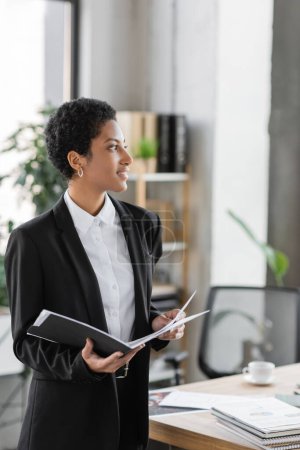 joyful african american businesswoman holding folder with papers and looking away in office