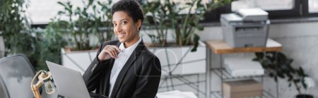pleased african american businesswoman in black blazer holding laptop and smiling at camera in contemporary office, banner