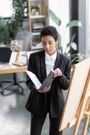 Foto de Mujer de negocios afroamericana en traje negro mirando documentos en carpeta mientras está de pie cerca del tablero de notas en la oficina - Imagen libre de derechos