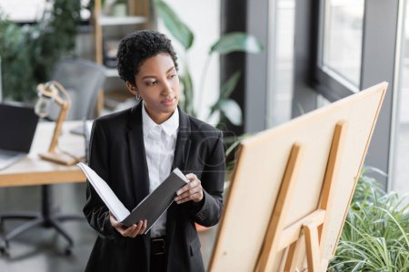 stylish african american businesswoman in black blazer holding folder and looking at note board in office