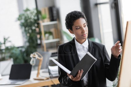 Foto de Elegante mujer de negocios afroamericana sosteniendo documentos y señalando borrosa pizarra en la oficina - Imagen libre de derechos