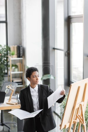 young african american businesswoman holding papers and looking at note board while working in office