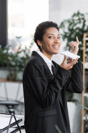 happy african american businesswoman pointing up with finger while recording voice message on smartphone in office