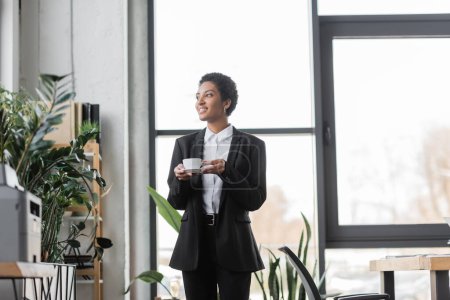 Téléchargez les photos : Femme d'affaires afro-américaine réussie en costume noir regardant loin tout en se tenant avec tasse de café dans le bureau contemporain - en image libre de droit