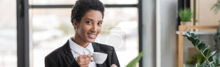 Téléchargez les photos : Joyeuse femme d'affaires afro-américaine en blazer noir tenant une tasse de café et regardant la caméra dans le bureau, bannière - en image libre de droit