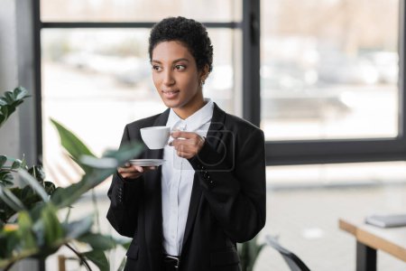 Téléchargez les photos : Femme d'affaires afro-américaine souriante dans un blazer noir debout avec une tasse de café et regardant loin dans le bureau - en image libre de droit