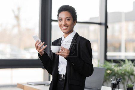 Téléchargez les photos : Heureuse femme d'affaires afro-américaine avec téléphone portable et tasse de café en regardant la caméra dans le bureau - en image libre de droit