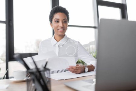 happy african american businesswoman looking at blurred laptop while working with papers in office