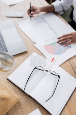 high angle view of eyeglasses and blank notebook near cropped african american businesswoman working with infographics in office