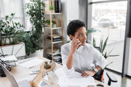 pensive african american businesswoman sitting near documents and laptop while looking away in office