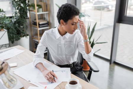 Foto de Mujer de negocios afroamericana cansada sentada cerca de documentos con cartas y sufriendo de dolor de cabeza en la oficina - Imagen libre de derechos