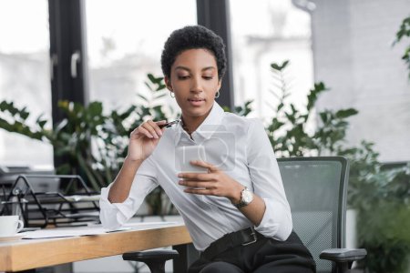 african american businesswoman holding pen and using cellphone while sitting at workplace in office