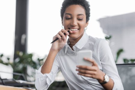 Téléchargez les photos : Joyeuse femme d'affaires afro-américaine en chemisier blanc tenant un stylo et regardant le téléphone mobile dans le bureau - en image libre de droit