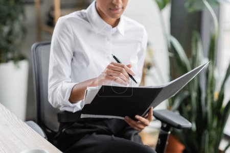 Teilaufnahme einer afrikanisch-amerikanischen Geschäftsfrau in weißer Bluse, die mit Stift und Mappe mit Dokumenten im Büro sitzt