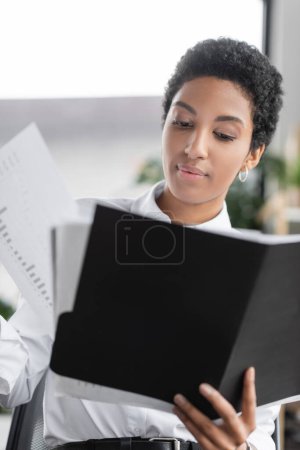 african american businesswoman holding folder and looking at papers with charts in office
