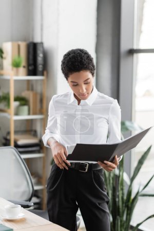 stylish african american businesswoman in white blouse and black pants standing with folder in office