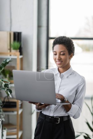 Téléchargez les photos : Femme d'affaires afro-américaine gaie utilisant un ordinateur portable tout en se tenant dans le bureau moderne - en image libre de droit