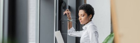 Photo for Side view of young african american businesswoman in white blouse holding laptop near windows in office, banner - Royalty Free Image