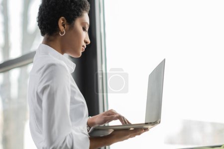 Foto de Vista lateral de la mujer de negocios afroamericana en blusa blanca de pie cerca de la ventana en la oficina y utilizando el ordenador portátil - Imagen libre de derechos