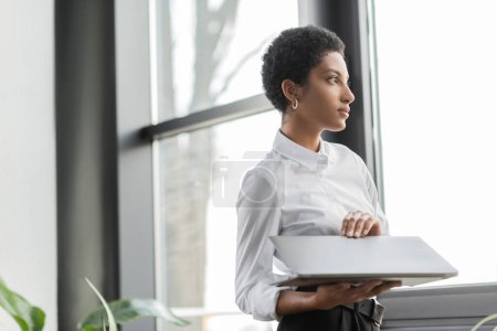 pensive african american businesswoman holding laptop and looking away near window in office