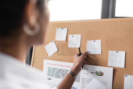 Téléchargez les photos : Vue recadrée de femme d'affaires afro-américaine toucher note de papier avec infographies sur carton de liège au bureau - en image libre de droit