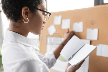 Foto de Vista lateral de la mujer de negocios afroamericana de pie cerca del tablero de notas borrosas y la celebración de cuaderno en blanco en la oficina - Imagen libre de derechos